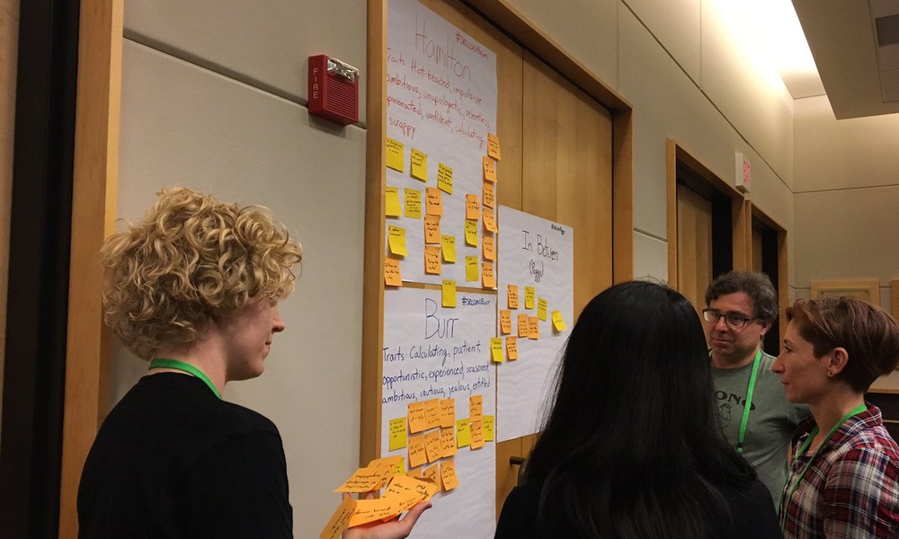 A woman with curly blonde hair holds sticky notes for people gathered around, sorting the sticky notes into groups.